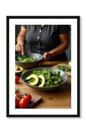 A plus-size black woman is preparing a green salad with avocado slices on a wooden table. The background shows a bowl of cherry tomatoes and a cutting board with herbs