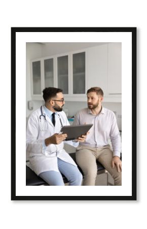 Serious Latin general practitioner man asking male patient about health complaints, listening, holding pad with medical records, history, giving consultation in clinic examination room