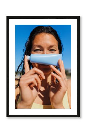 Woman holding sunscreen tube in front of face on sunny beach
