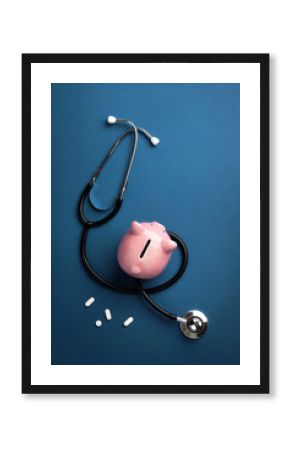 A stethoscope with pink piggy bank and with pills on blue background. Top view, flat lay.