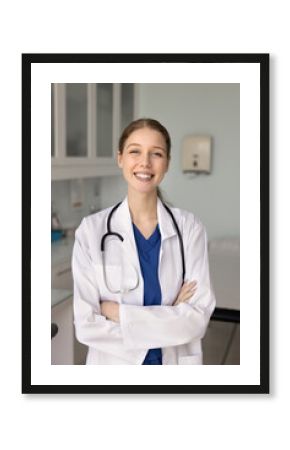 Happy beautiful young physician woman in lab coat posing for profile picture in doctor office, standing with hands folded in confident gesture, looking at camera with toothy smile. Vertical portrait