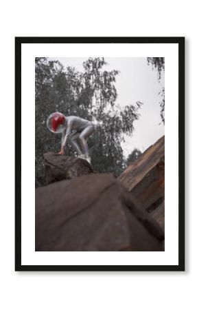 Low angle of positive young female with dyed red hair wearing silver space suit and glass helmet and looking away while walking on rocky formation