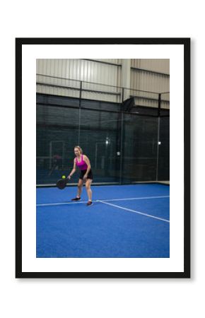 Woman wearing pink top holding padel racket, focusing on ball on padel court, copy space