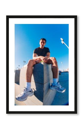 A young man with short curly hair sits on a concrete bench in an urban outdoor space under a bright blue sky, wearing a black t-shirt, yellow shorts, and white sneakers, with a relaxed posture