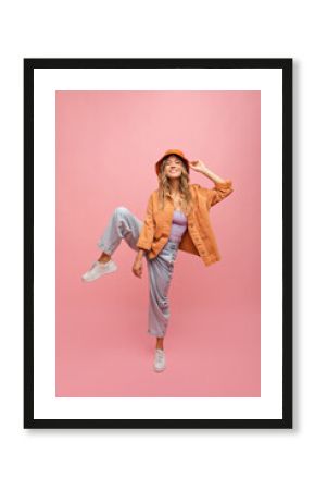 Full-length, amusing young fair-skinned girl stands with her leg raised against background of pink wall with space for text. Blonde with wavy, loose hair wears lilac top, orange jacket and jeans.