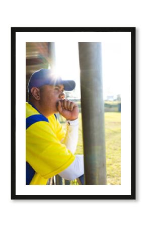 Sitting in dugout at chain-link fence, Hispanic man wearing yellow jersey, blue cap, copy space
