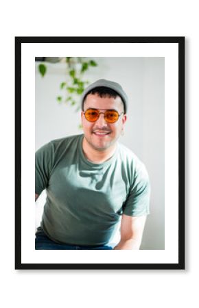 A man in his thirties sits casually with sunglasses and a hat, smiling in a bright space