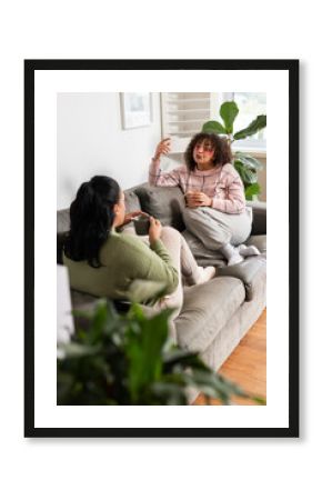 Mother and daughter chatting on sofa at home holding ceramic mugs beside potted plant, copy space