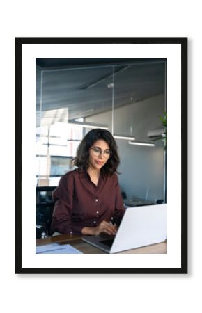 Young professional it specialist latin hispanic business lady working on laptop pc sitting at desk in modern office space. Middle eastern indian woman using computer technology app for work. Vertical