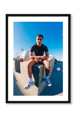 A young man with short curly hair sits on a concrete bench in an urban outdoor space under a bright blue sky, wearing a black t-shirt, yellow shorts, and white sneakers, with a relaxed posture