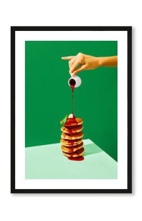 Food pop art photography. Close up. Female hand pouring sweet jam on delicious pancakes with butter over green background. Complementary colors.