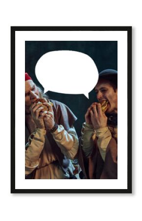 Two men, medieval pirates in hat and bandana, passionately eating burgers against dark vintage background with abstract speech bubble. Concept of food, adventure, comparison of eras, leisure