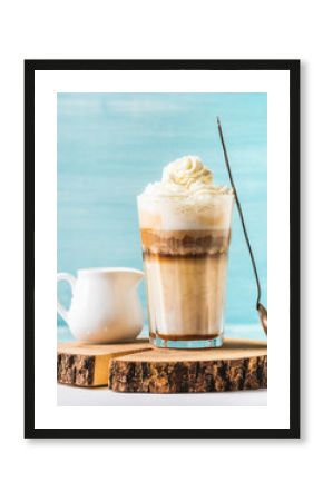 Latte macchiato with whipped cream, serving silver spoon and white pitcher on wooden round board over blue painted wall background