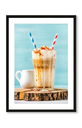 Latte macchiato with whipped cream and caramel sauce in tall glass with straws served with pitcher on wooden board over blue painted wall background, selective focus, vertical composition