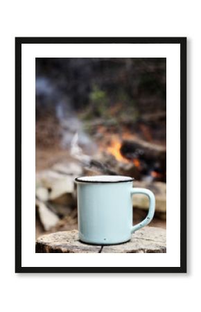 Blue enamel cup of hot steaming coffee sitting on an old log by an outdoor campfire. Extreme shallow depth of field with selective focus on mug.