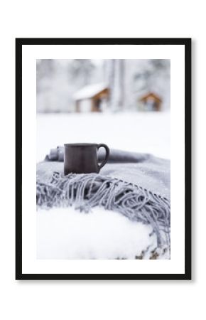 cup with hot coffee on a gray scarf on a snow-covered table in a winter forest next to wooden houses