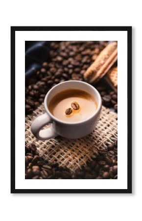       Cup of coffee in the middle of coffee beans with biscuits and tablecloth. Grained product. Hot drink. Close up. Harvesting. Natural background. Energy. Fresh espresso with beans on the foam.