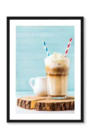 Latte macchiato with whipped cream in tall glass with two straws and pitcher on wooden board over blue painted wall background, selective focus, copy space, vertical composition
