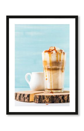 Latte macchiato with whipped cream and caramel sauce in tall glass on wooden board over blue painted wall background, selective focus, copy space, vertical composition