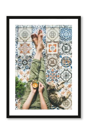 Lazy morning on terrace. Flat-lay of womans legs in cosy linen pants, plant, cat and cup of coffee in hand over colorful moroccan tile floor, top view
