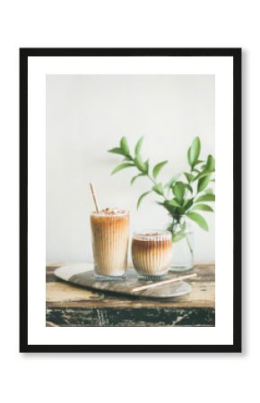 Iced coffee in tall glasses with milk and straws on board, white wall and green plant branches at background, copy space. Summer refreshing beverage ice coffee drink concept