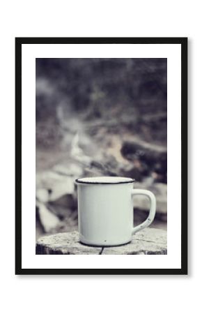Cup of hot steaming coffee sitting on an old log by an outdoor campfire with a vintage antique edit. Extreme shallow depth of field with selective focus on mug.