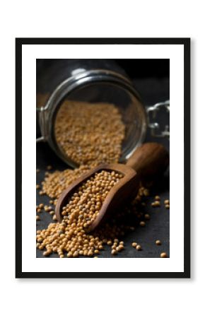 Heap of raw, unprocessed mustard seed kernels in glass jar and wooden scoop on black table