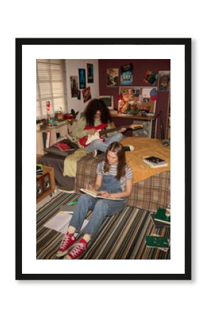 Two teenagers engaging in activities at cozy bedroom with posters and retro study materials. One teenager reading a book while the other playing a red electric guitar on the bed