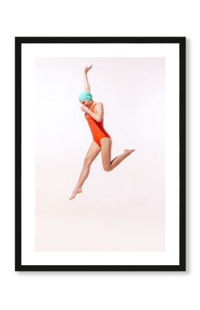 Portrait of young woman in swimming suit and cap jumping into water isolated over grey studio background