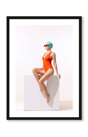 Portrait of young stylish woman in red swimming suit and sunglasses sitting, posing isolated over grey studio background