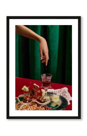 Female hand with wine drops over wine glass, surrounded by split pomegranate, pearls, and flowers on silver tray against green velvet background. Concept of party, celebration, high-society