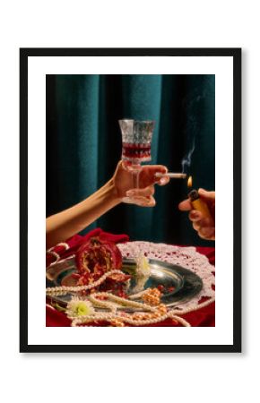 Female hand holding wine glass and cigarette, surrounded by split pomegranate, pearls, and white flowers on silver tray against green velvet background. Concept of party, celebration, high-society